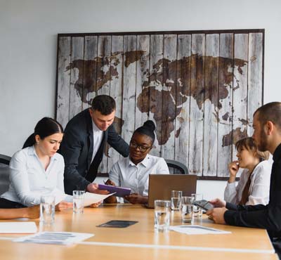 Five colleagues sitting in a meeting room looking at handouts and a tablet. There is an art piece hanging on the wall of the map of the world.